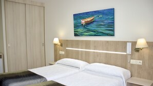 Habitación de hotel con cabecera de madera, dos camas con ropa blanca, un cuadro de un barco en el agua y lámparas de noche.