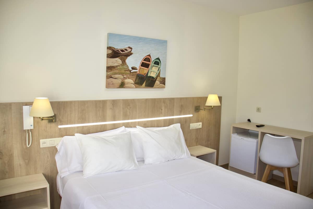 Una habitación de hotel con cama, mesitas de noche, un escritorio con silla, un refrigerador pequeño y un cuadro en la pared.