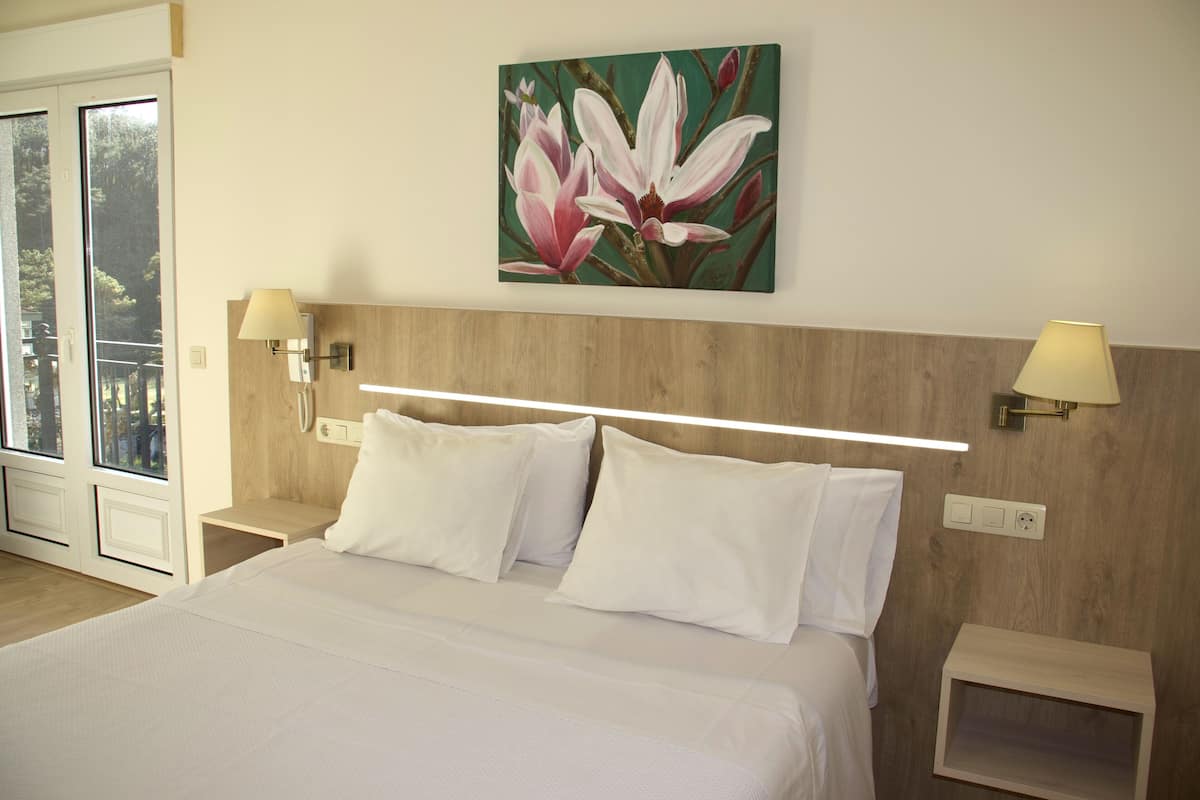 Habitación de hotel con cama, dos lámparas de noche, un cuadro de flores en la pared y ventana con vista a árboles.