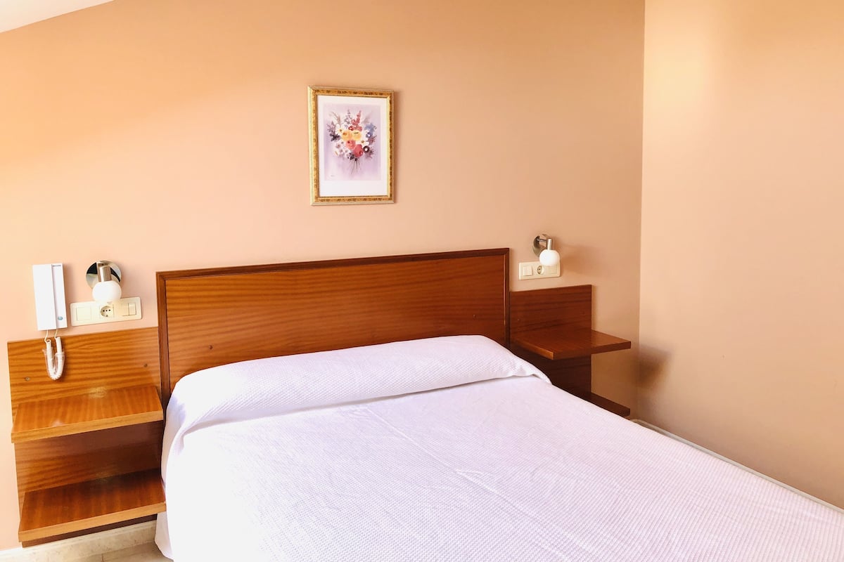 Habitación de hotel con cabecera de madera, colcha blanca, mesita de noche con lámpara y un cuadro con motivos florales en la pared.