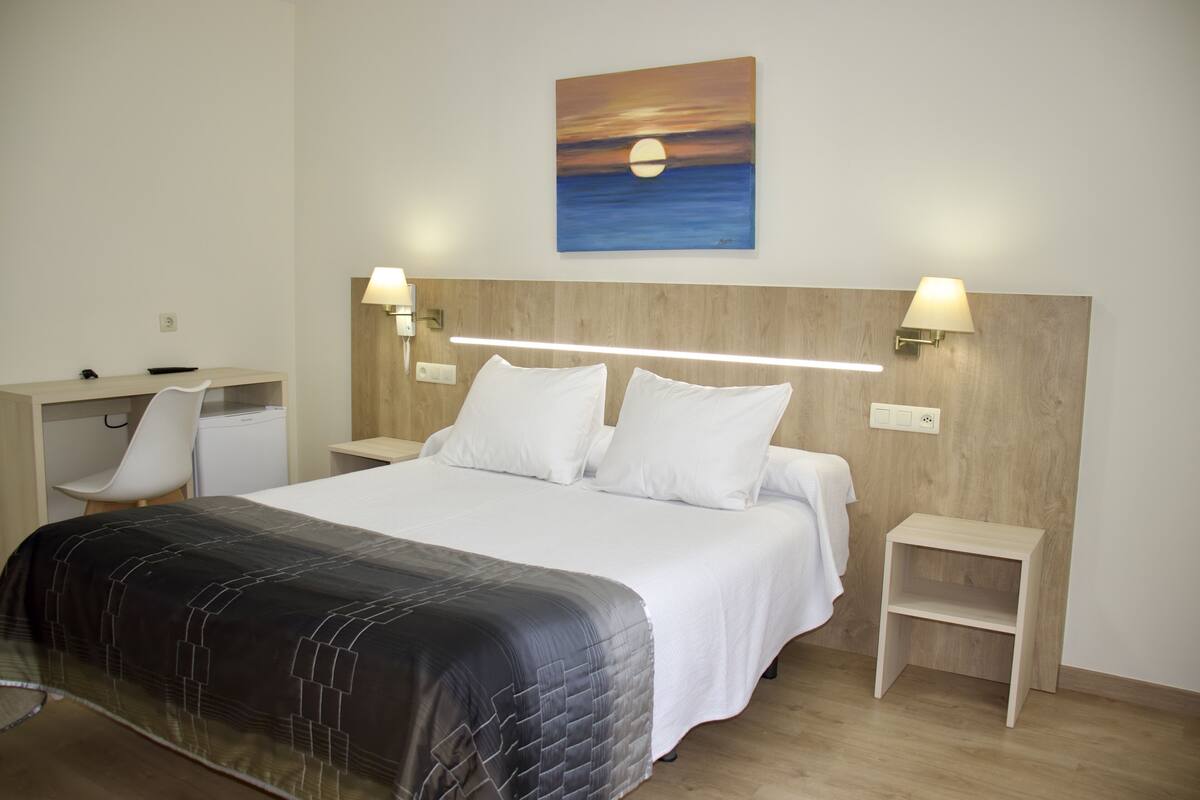 Una habitación de hotel con cama, mesitas de noche, un escritorio, una silla y un cuadro con una puesta de sol en la pared.