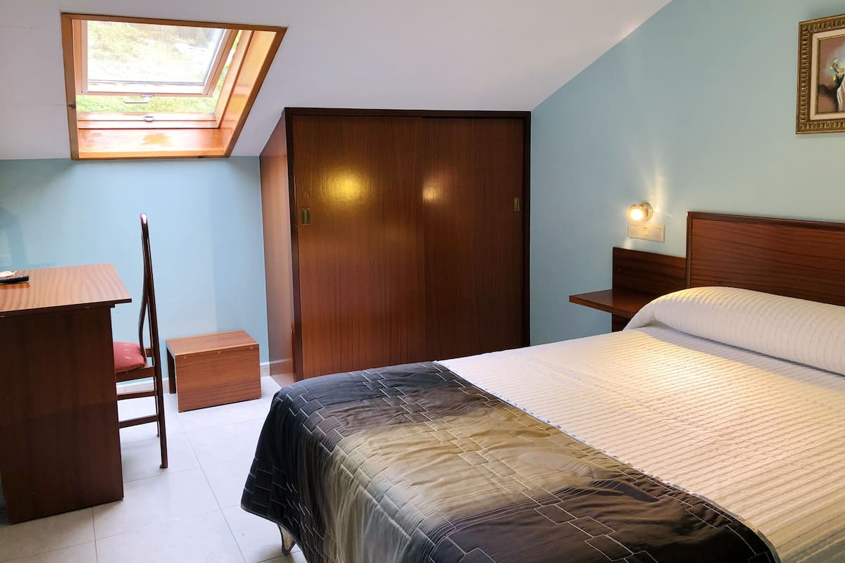Un dormitorio con cama, escritorio, silla, armario, tragaluz y mesita de noche con lámpara.
