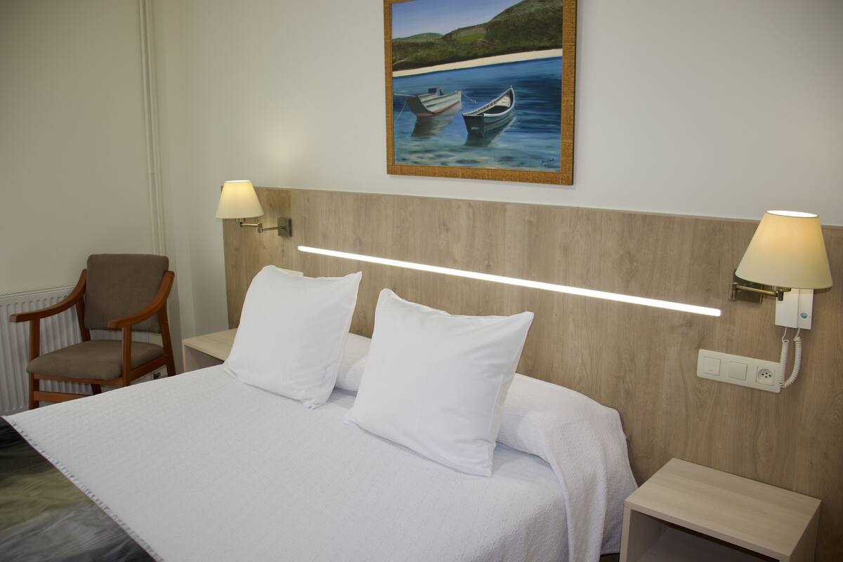 Una cama bien hecha con sábanas blancas, dos lámparas de noche, un cabecero de madera, una silla y un cuadro con barcos en la pared.