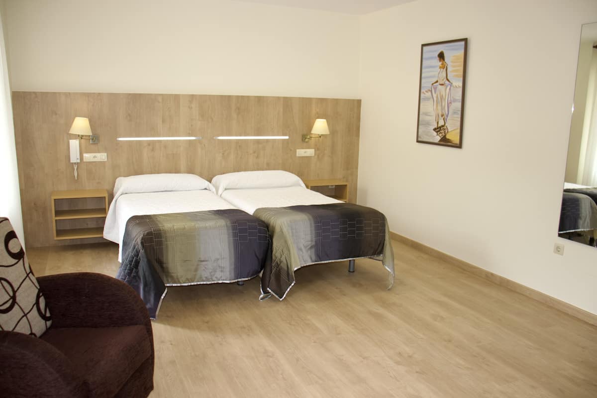 Habitación de hotel con dos camas, una mesita de noche, una silla y un cuadro en la pared.