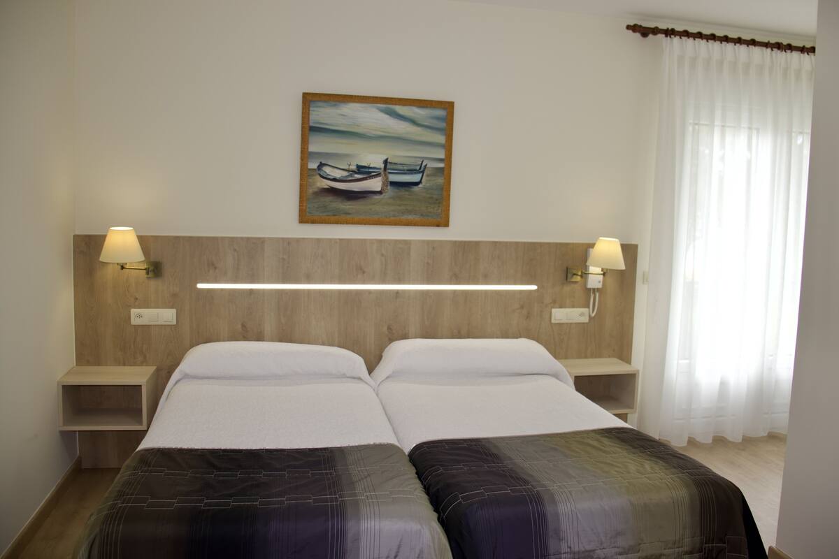Habitación de hotel con dos camas, un cuadro de barcos en la pared y una mesita de noche con una lámpara.