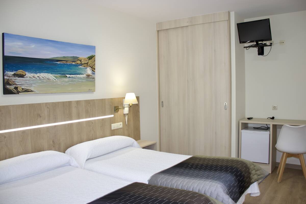 Habitación de hotel con dos camas, un escritorio con silla, un televisor montado en la pared y un cuadro de una playa.