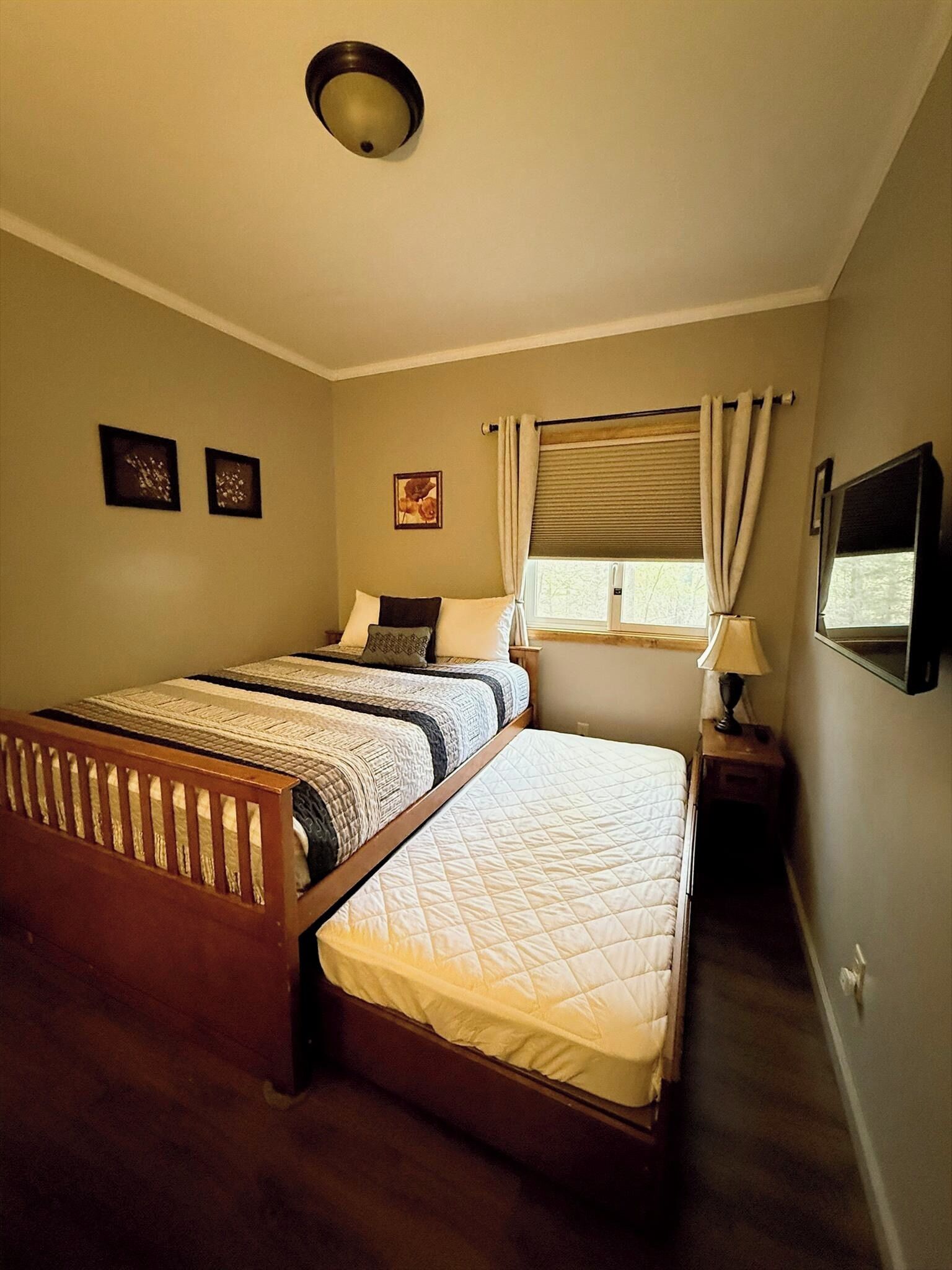 Une chambre avec un lit double, un lit simple, une fenêtre avec des rideaux, une table de chevet avec une lampe et un téléviseur fixé au mur.