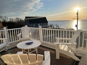 A deck with white chairs and a table, overlooking a body of water and a house.