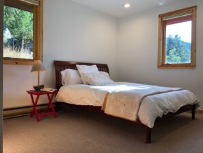 Une chambre à coucher avec un lit en bois, du linge de lit blanc, une table de chevet rouge, une lampe et deux fenêtres laissant entrer la lumière naturelle.