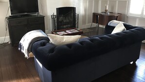Ett vardagsrum med en mörkblå soffa, en öppen spis, en tv som står på ett skåp och ett litet bord med böcker.