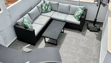 Un área de estar exterior moderna con un sofá seccional gris, una mesita negra y una mesita auxiliar de mimbre.