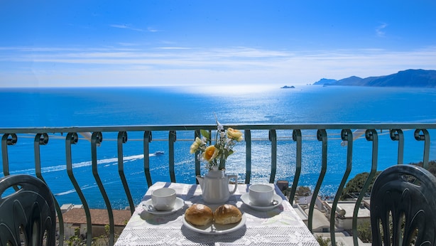 Une table recouverte d’une nappe blanche, deux tasses, deux petits pains et un vase rempli de fleurs, le tout disposé devant un paysage marin pittoresque.