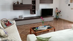 Uma sala de estar moderna com televisão, prateleiras decoradas, uma mesa de centro com um vaso e um sofá com almofadas.