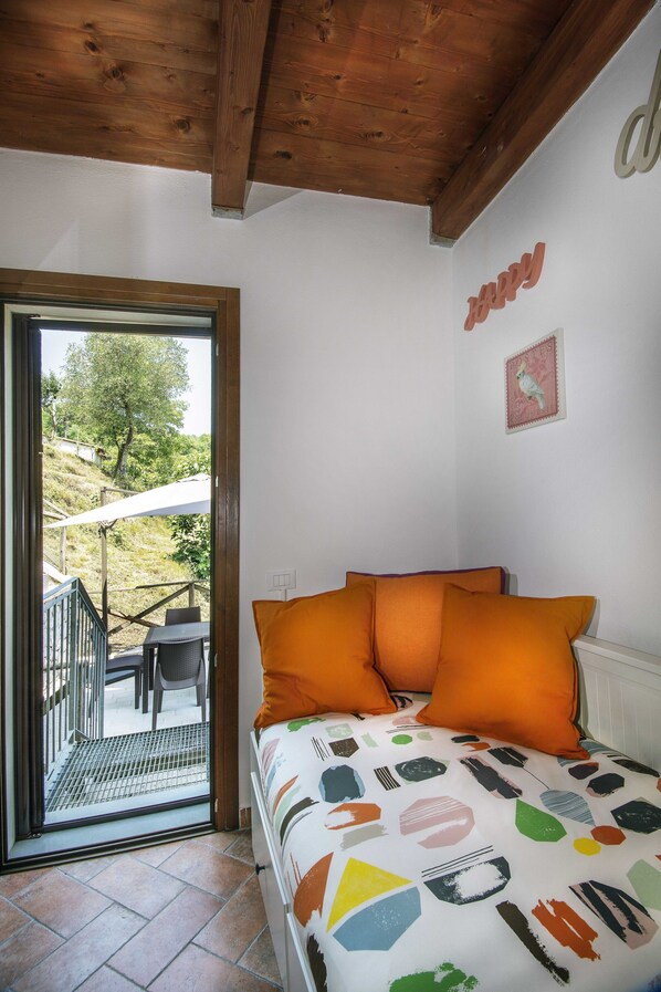 Une chambre avec un lit, des oreillers orange, un mur blanc orné d’un panneau indiquant le mot « joie », un plafond en bois et une porte donnant sur un espace extérieur avec une table et des chaises.