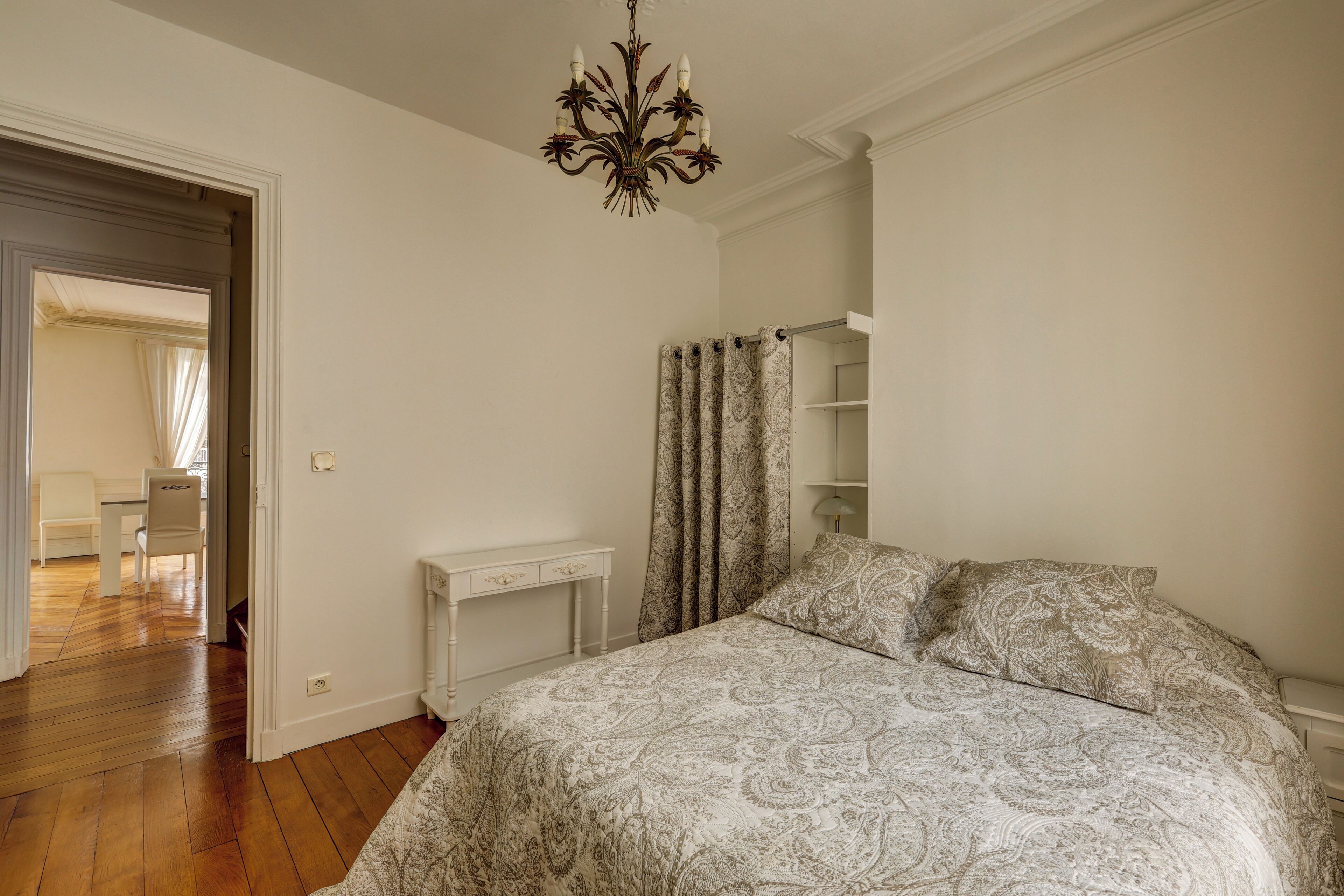 A bedroom with a patterned bedspread, a white bedside table, a chandelier, and a bathroom with a shower curtain.