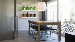 Een eethoek met een houten tafel en zwarte frame stoelen, een witte hanglamp en planken met groene en bruine flessen.