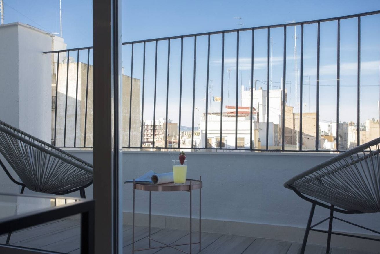 Een balkon met twee stoelen, een klein tafeltje met een drankje, en uitzicht op de stad.
