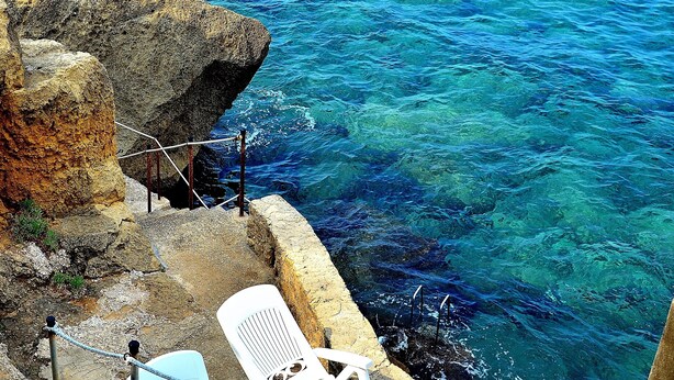 A rocky coastline with clear blue water, a stone wall, and two white chairs near the edge.