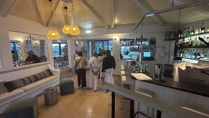 A modern bar area with a high ceiling, exposed beams, and a bar counter with various bottles and glasses. There are three hexagonal hanging lights, a trash can, and a seating area with a bench and cushions. - Alto Club, Alvor. Quiet, comfortable apartment in this acclaimed holiday resort (Alvor)