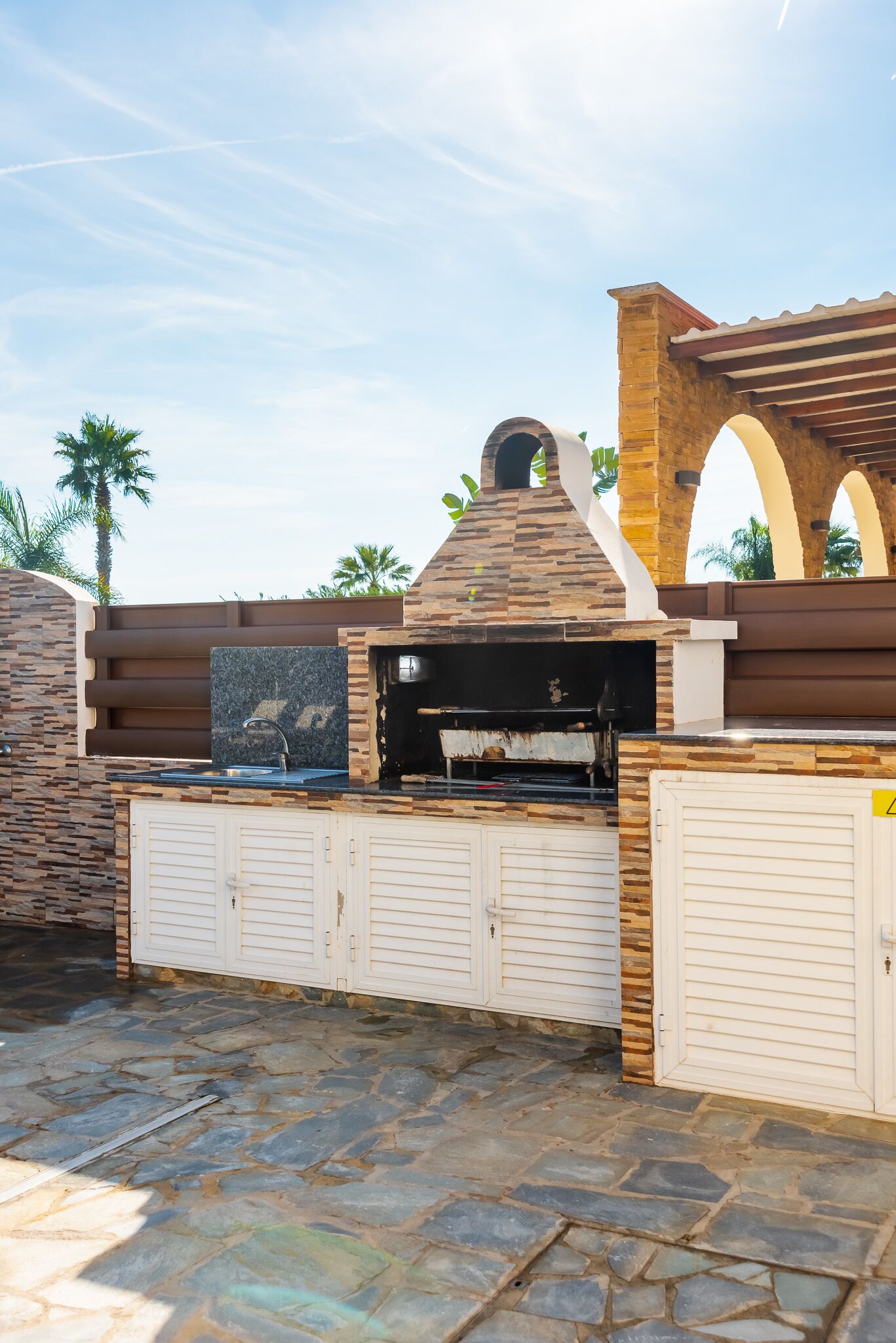 An outdoor barbecue area with a stone and brick structure, a grill, and a stone-tiled floor.