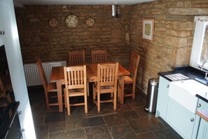 A dining area with a stone wall, wooden table, and chairs. There is a window with a curtain, a trash can, and a kitchen area with cabinets and a sink. - Spacious barn conversion in a tranquil Oxfordshire village (Shutford)