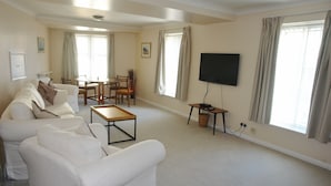 Ein Wohnzimmer mit einer Sofagarnitur, einem Couchtisch, einem an der Wand montierten Fernseher und einem Essbereich mit Tisch und Stühlen.