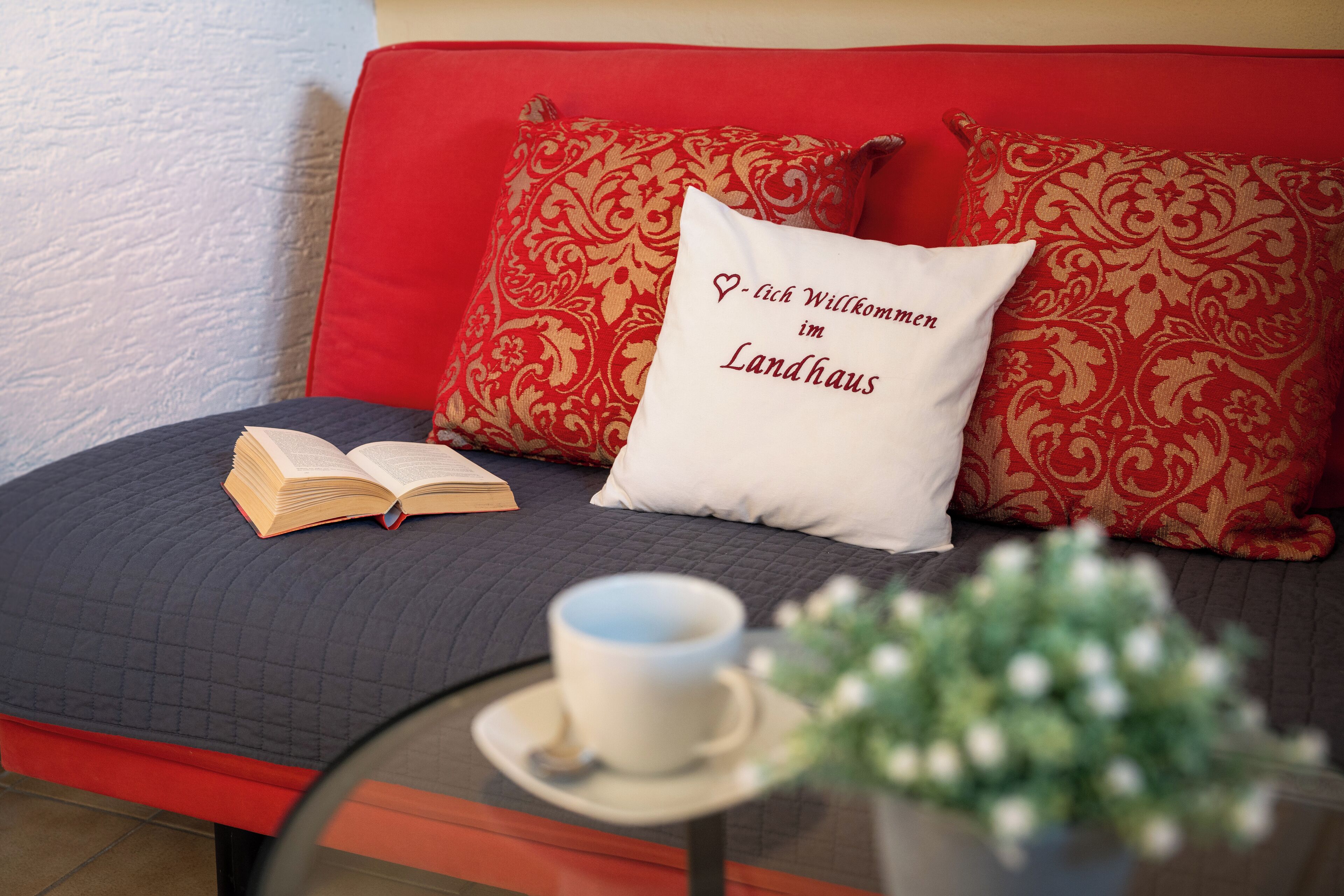 A cozy room with a red sofa, a white pillow with text, an open book, a cup on a saucer, and a small table with a plant.