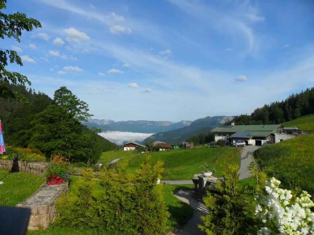 A rural landscape with a path leading to a building with a green roof, surrounded by greenery and mountains in the background.
