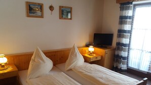 Een tweepersoonsbed met witte lakens, een nachtkastje met een lamp, een televisie op een houten kast en twee ingelijste foto's aan de muur.