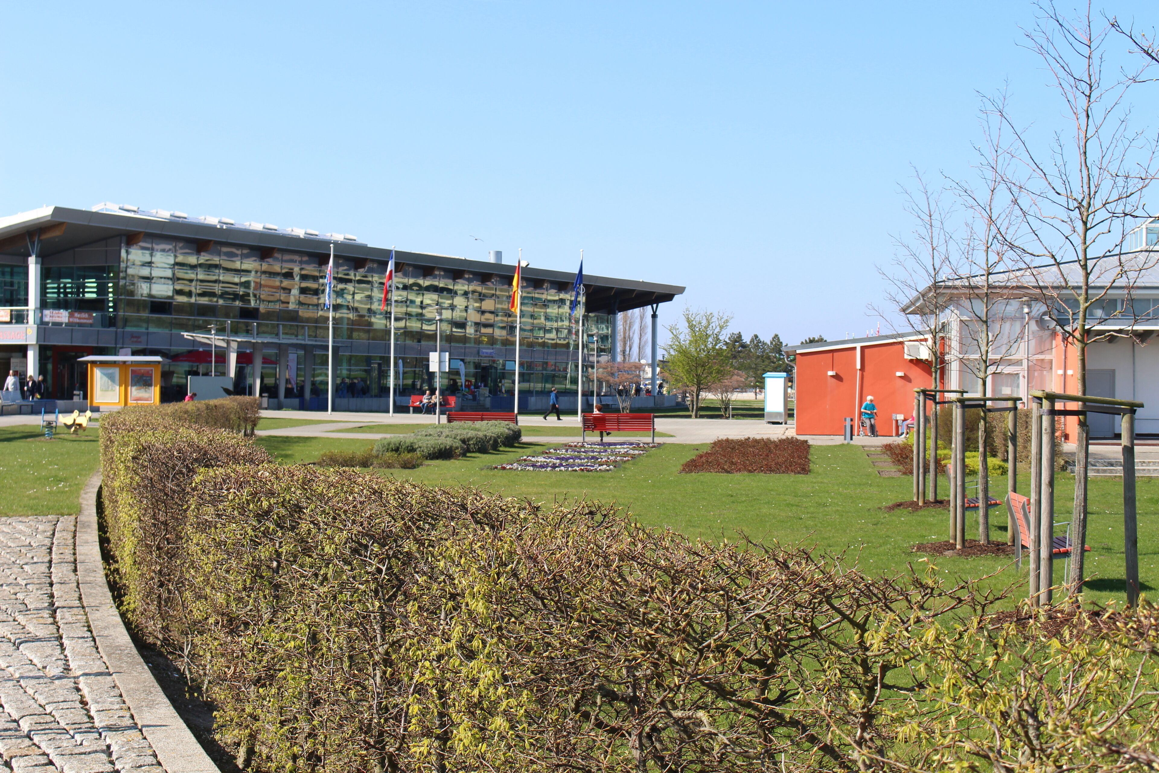 Ein modernes Gebäude mit Glasfassade, ein gepflegter Garten mit Bänken und ein klarer Himmel.