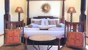 Un lit à baldaquin avec des serviettes roulées, deux chaises avec des coussins à motifs et une table ronde dans une chambre d’hôtel.