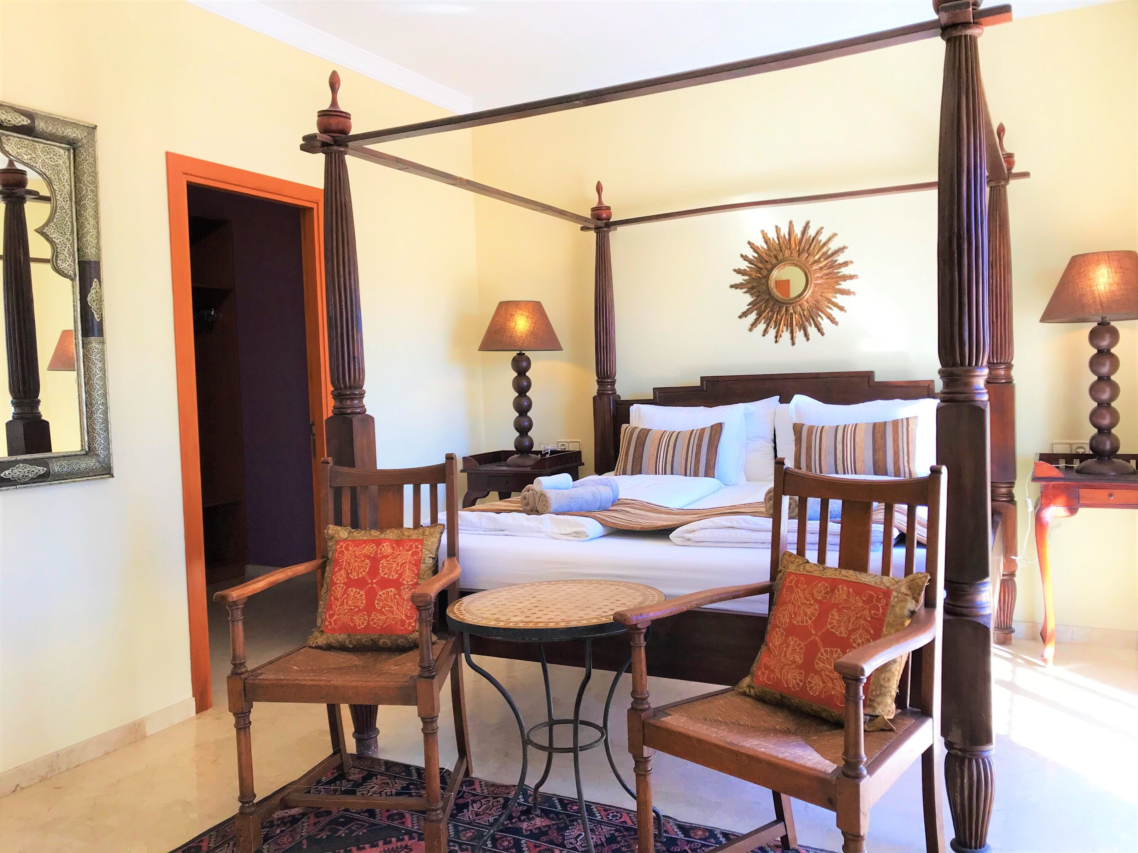 A four-poster bed with a sunburst mirror, two wooden chairs with red cushions, a small round table, and a patterned rug.