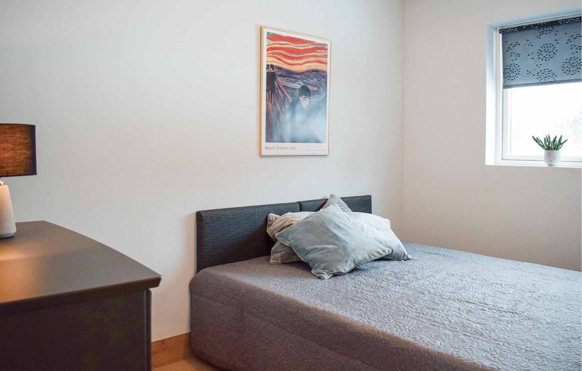 A bedroom with a bed, a nightstand, a lamp, a window with a plant, and a framed picture on the wall.