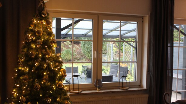 Un árbol de Navidad con luces y adornos junto a una ventana que da vista a muebles de exterior.