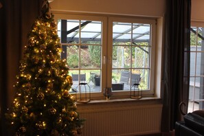 Un sapin de Noël décoré de guirlandes lumineuses et d’ornements, placé près d’une fenêtre donnant sur du mobilier d’extérieur.