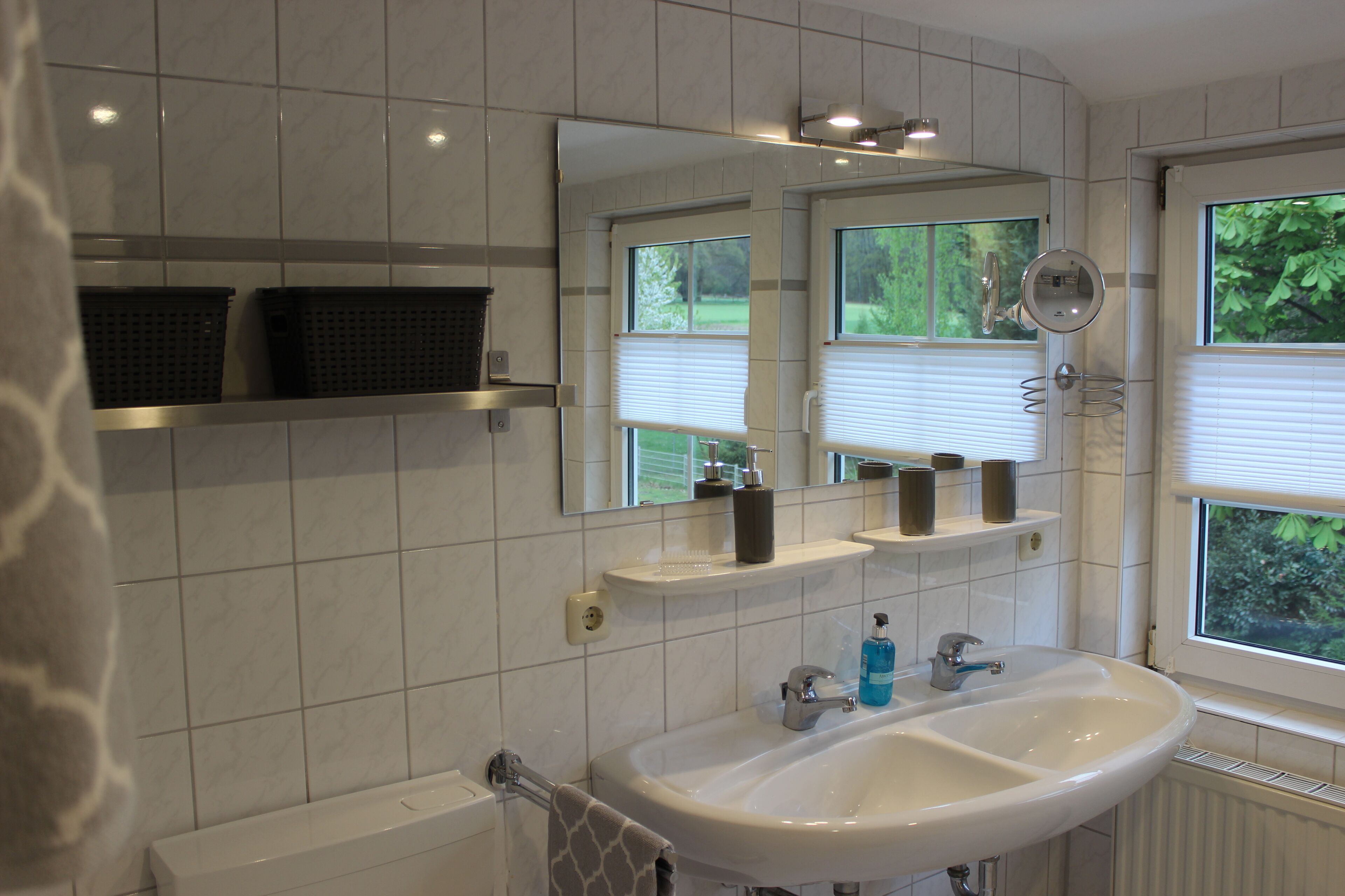 Ein Badezimmer mit einem weißen Waschbecken, zwei Spiegeln und einem Fenster mit Jalousien.