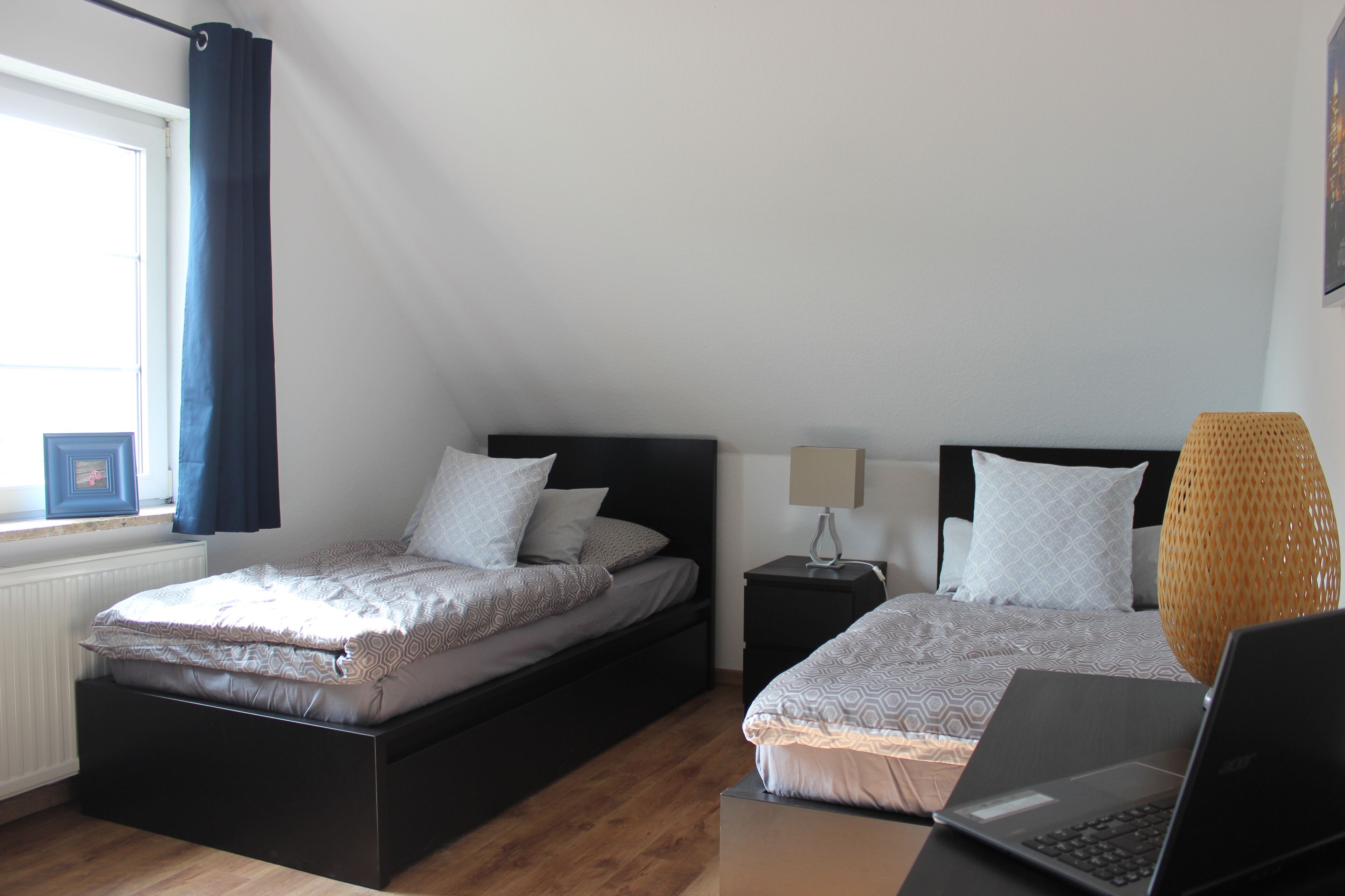 Ein Schlafzimmer mit zwei Betten, einem Fenster mit Vorhängen, einem Nachttisch mit Lampe und einem Laptop auf einem Ständer.