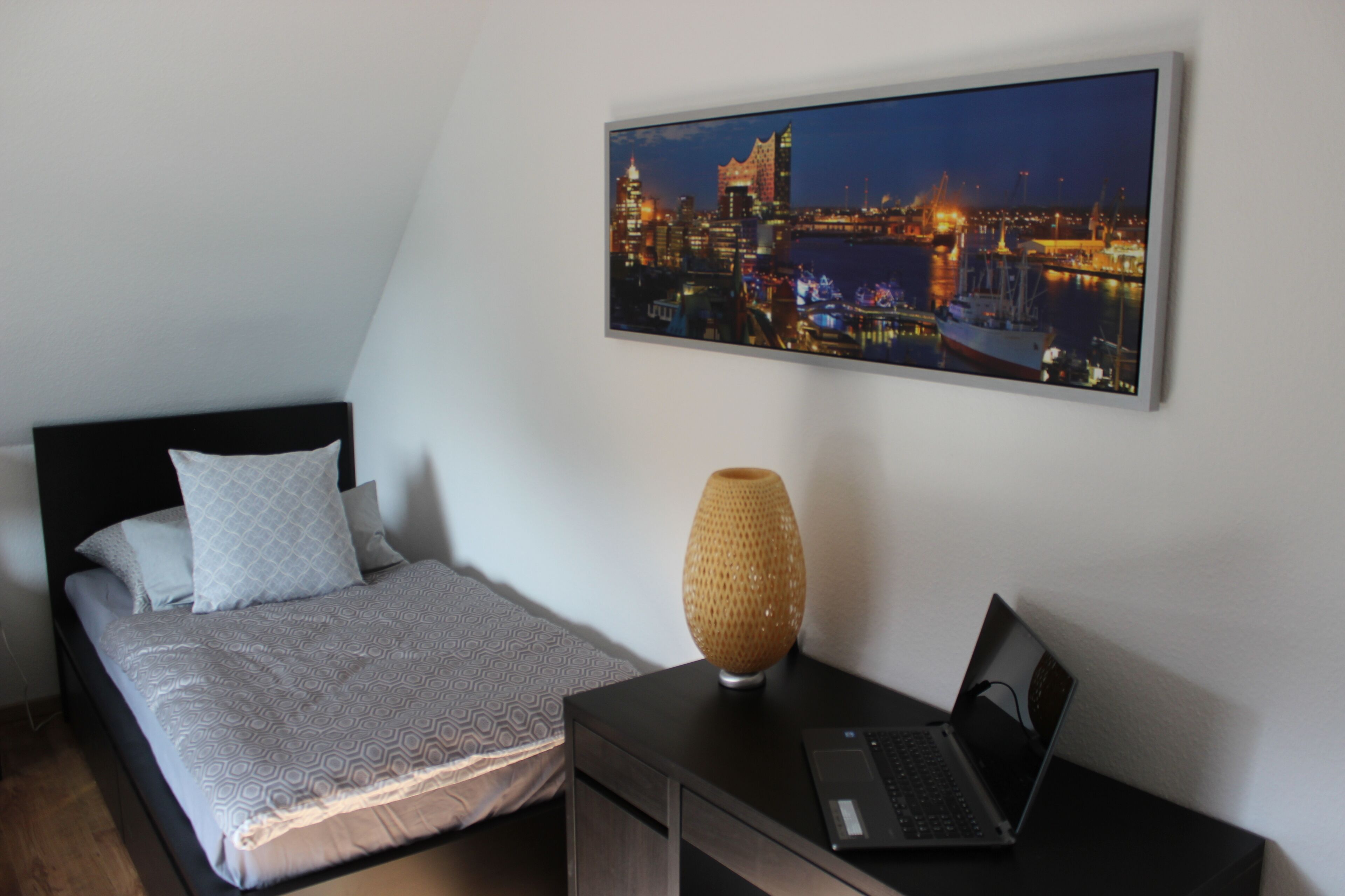 A bedroom with a bed, a nightstand, a laptop, a lamp, and a cityscape painting on the wall.