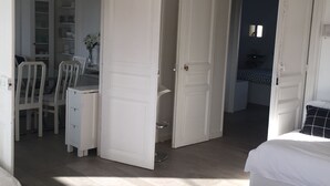 Een kamer met een open deur naar een eetgedeelte, een bed met een felroze kussen, en een kleine tafel met een glazen blad.