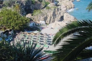 A coastal view with rocky cliffs, a beach with umbrellas, and palm trees.