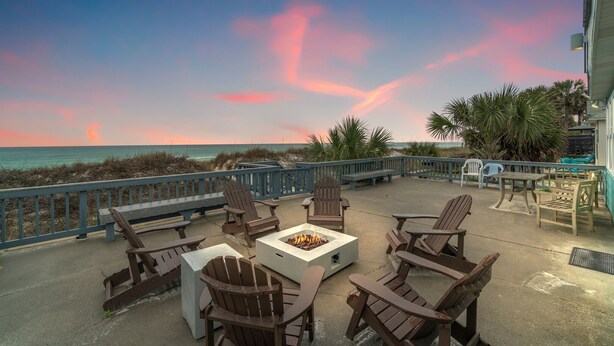 A patio with Adirondack chairs, a fire pit, and a view of the ocean at sunset.