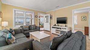 A living room with a grey sofa set, a coffee table, a flat-screen TV on a white entertainment unit, and a door leading to another room.