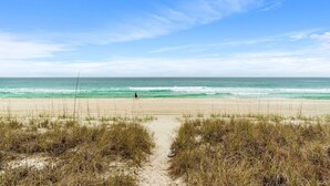 A beach with a clear sky, a sandy path leading to the water, and a person walking.