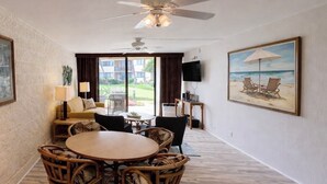 A hotel room with a dining area, a ceiling fan, a TV, and a painting of a beach scene.