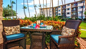 A wicker table set with a glass top, surrounded by wicker chairs with blue cushions, in front of a building with balconies and palm trees.