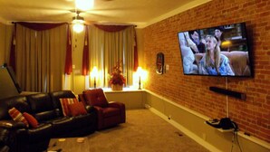 A living room with a brick wall, a television mounted on the wall, a ceiling fan, and a leather sofa with red cushions. - DOWNTOWN INDY HOME IN HISTORIC LOCKERBIE SQUARE  - 7 BLOCKS FROM CITY CENTER! (Indianapolis)