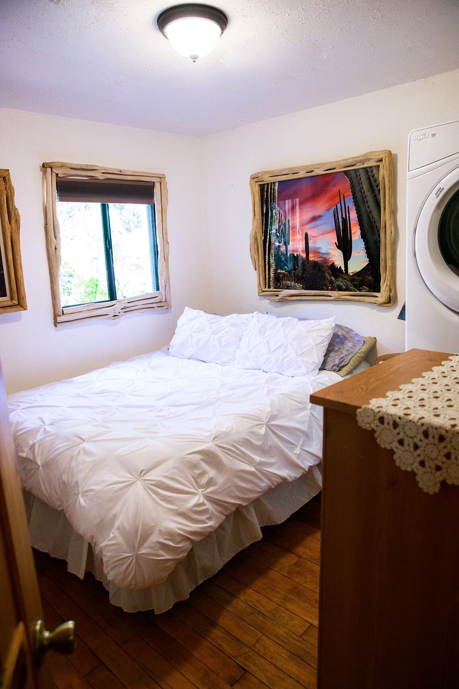 A bedroom with a bed, a window, a painting, a washing machine, and a wooden dresser.