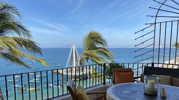 A balcony with a view of the sea, a sailboat, and a dining area with chairs and a table.