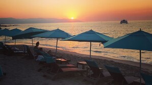 A beach with umbrellas and lounge chairs, a boat in the water, and a sunset in the background.