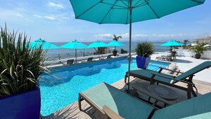 A poolside area with lounge chairs and umbrellas, a clear view of the ocean, and a tiled pool.